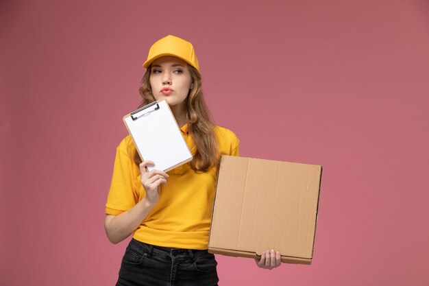 Front view young female courier in yellow uniform holding notepad and food delivery package on pink background job uniform delivery color service worker