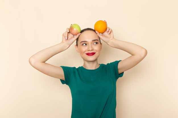 Free photo front view young female in dark green shirt and blue jeans holding apple and orange with smile on her face on beige