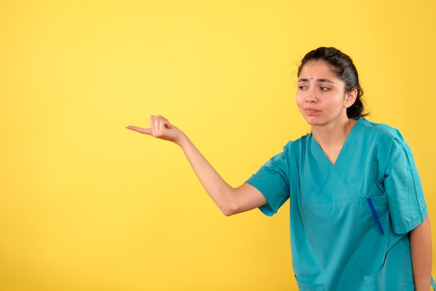 Front view of young female doctor in uniform pointing at left direction standing on yellow wall