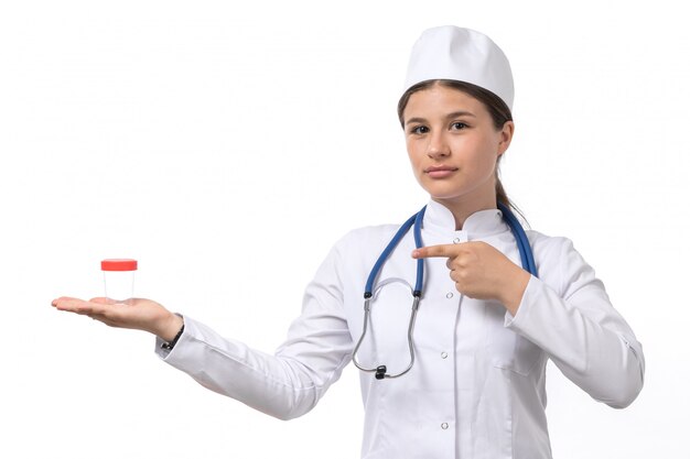 A front view young female doctor in white medical suit and white cap with blue stethoscope holding flask 