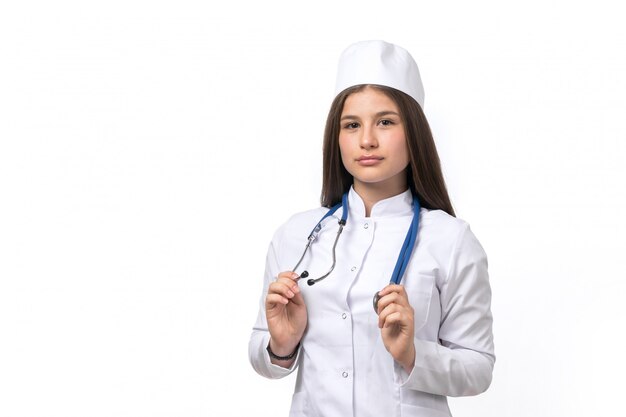 A front view young female doctor in white medical suit and white cap with blue stethoscope posing 