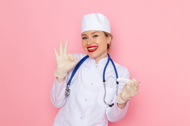 Front view young female doctor in white medical suit with blue stethoscope holding device smiling on the pink space medicine medical hospital