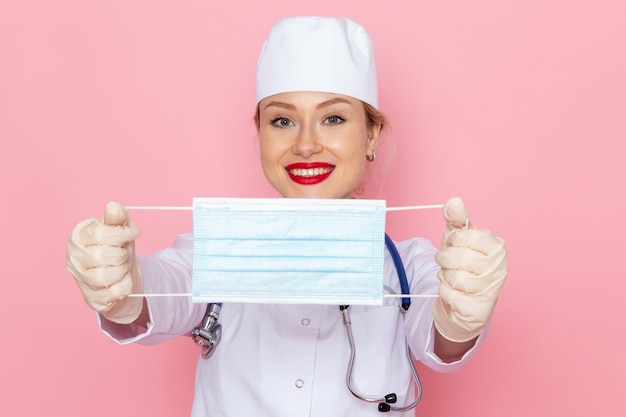 Front view young female doctor in white medical suit with blue stethoscope holding sterile mask on the pink space nurse