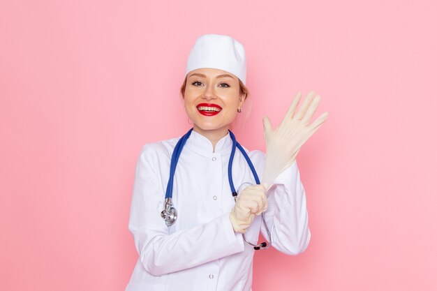 Front view young female doctor in white medical suit with blue stethoscope smiling and wearing gloves on the pink space medicine medical hospital