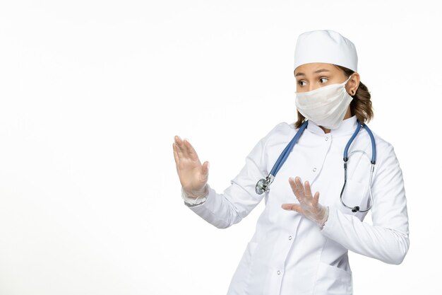 Front view young female doctor in white medical suit and with mask due to coronavirus on light-white surface