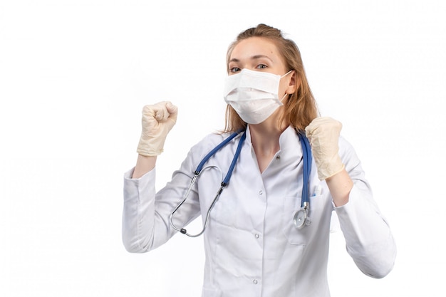 Free photo a front view young female doctor in white medical suit with stethoscope wearing white protective mask in gloves on the white