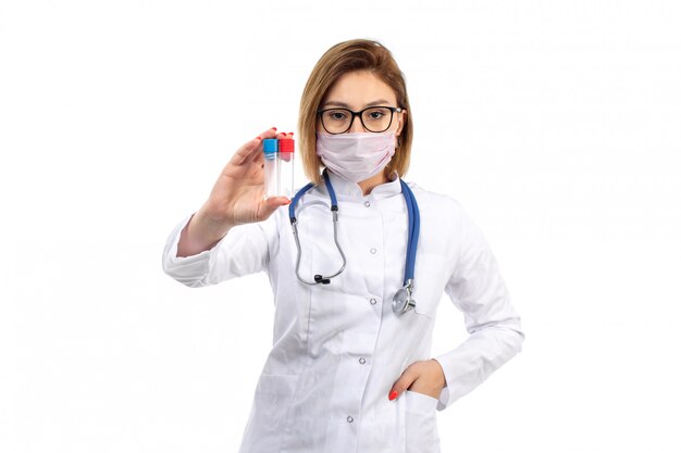 A front view young female doctor in white medical suit with stethoscope wearing white protective mask holding flasks on the white