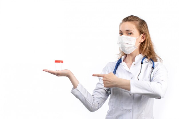 A front view young female doctor in white medical suit with stethoscope wearing white protective mask posing holding flask on the white