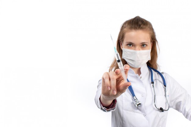 A front view young female doctor in white medical suit with stethoscope wearing white protective mask posing holding injection on the white