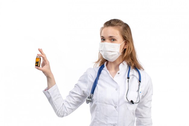 A front view young female doctor in white medical suit with stethoscope wearing white protective mask posing holding pills on the white