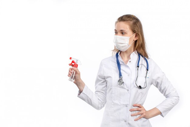 A front view young female doctor in white medical suit with stethoscope wearing white protective mask posing holding spray on the white