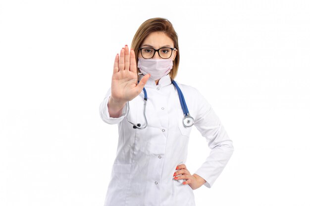 A front view young female doctor in white medical suit with stethoscope wearing white protective mask showing stop sign on the white