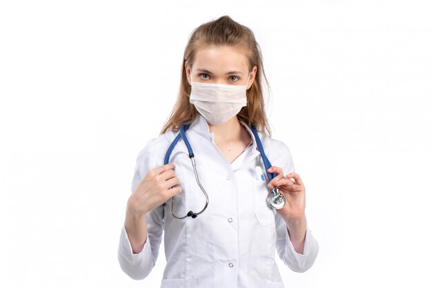 A front view young female doctor in white medical suit with stethoscope wearing white protective mask on the white