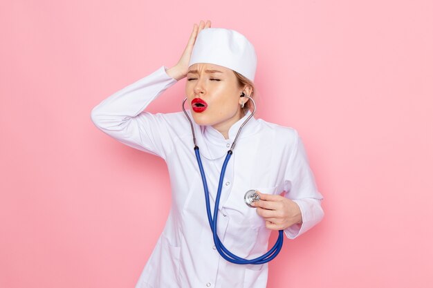 Front view young female doctor in white suit with blue stethoscope posing holding her head on the pink space  female work