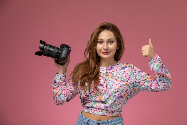 Front view young female in flower designed shirt and blue jeans holding black photo camera on the pink background 