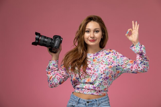 Front view young female in flower designed shirt and blue jeans holding camera smiling slightly on pink background 