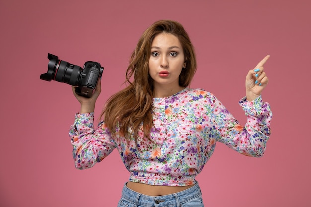 Front view young female in flower designed shirt and blue jeans taking a photo with camera on pink background 