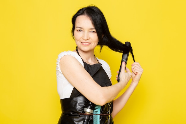 A front view young female hairdresser in white t-shirt black cape fixing making up her hair with tool smiling