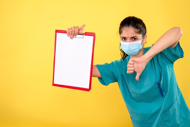 Front view of young female holding clipboard making thumb down sign on yellow wall