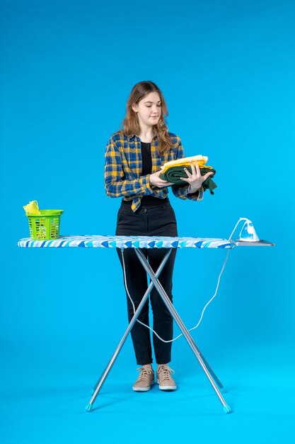 Front view of young female holding the folded clothes and standing behind the ironing board in the laundry room