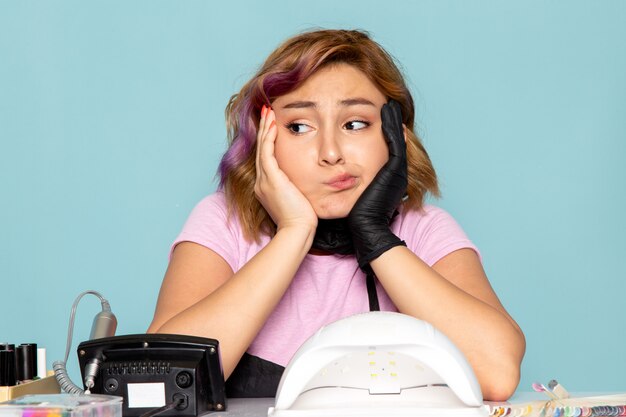 A front view young female manicure in pink t-shirt with black gloves and black mask getting bored on blue