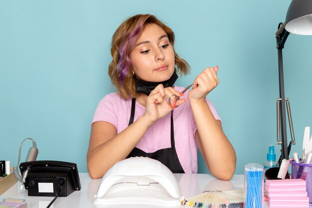 A front view young female manicure in pink t-shirt with black gloves and black mask sitting in front of the table fixing her nails on blue