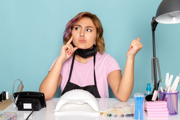 A front view young female manicure in pink t-shirt with black gloves and black mask sitting in front of the table and thinking on blue