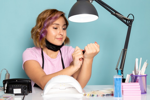 A front view young female manicure in pink t-shirt with black gloves and black mask sitting in front of the table working with her nails on blue