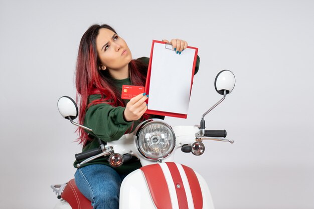 Front view young female on motorcycle holding note and bank card on a white wall