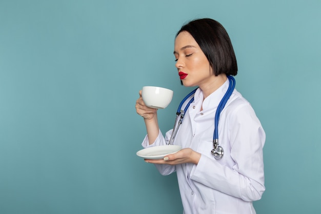 A front view young female nurse in white medical suit and blue stethoscope drinking tea