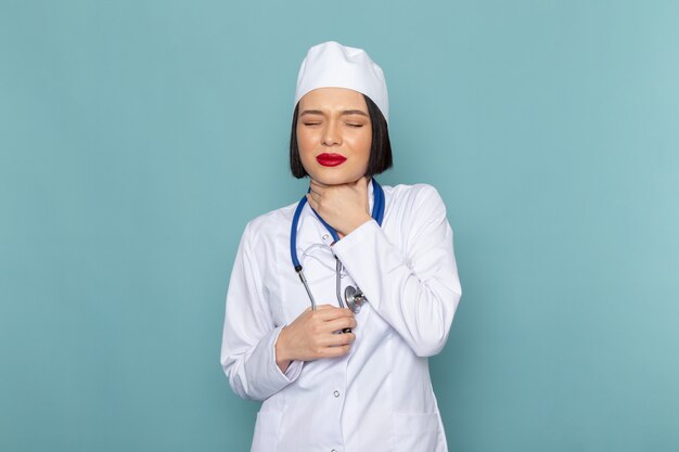 A front view young female nurse in white medical suit and blue stethoscope having throat issues