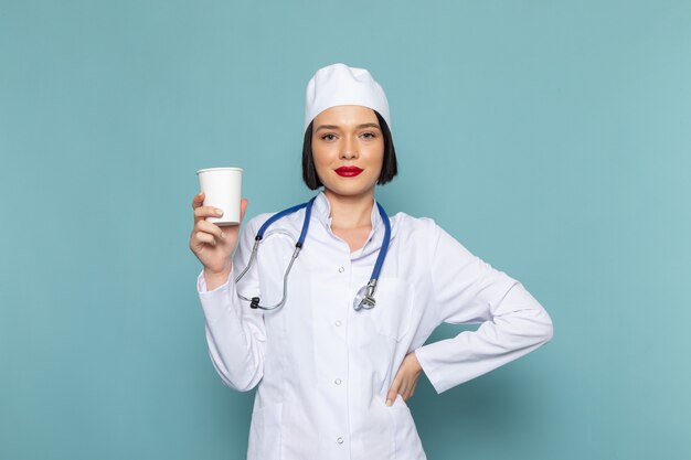 A front view young female nurse in white medical suit and blue stethoscope holding plastic glass on the blue desk medicine hospital doctor