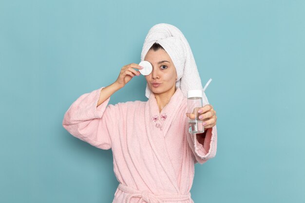 Front view young female in pink bathrobe cleaning her face from make-up on the blue wall shower cleaning beauty selfcare cream