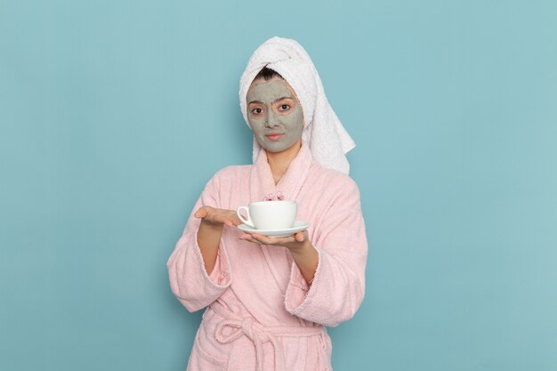 Front view young female in pink bathrobe holding cup of coffee on blue wall cleaning beauty selfcare cream shower