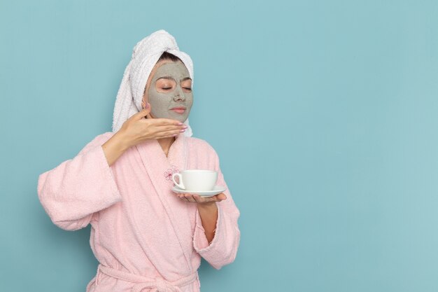 Front view young female in pink bathrobe with mask on her face holding coffee on the blue wall shower cleaning beauty selfcare cream