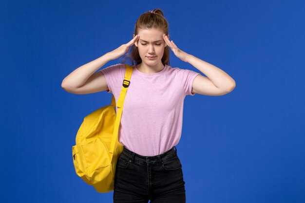 Front view of young female in pink t-shirt wearing yellow backpack having headache on blue wall