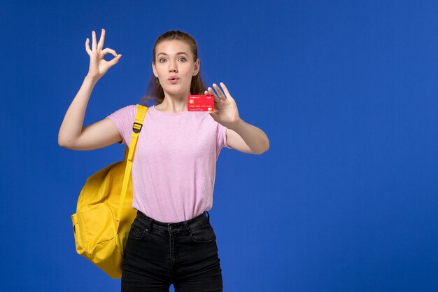 Front view of young female in pink t-shirt wearing yellow backpack holding plastic red card on the blue wall