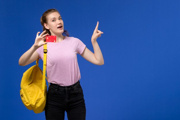 Front view of young female in pink t-shirt wearing yellow backpack holding plastic red card on the blue wall