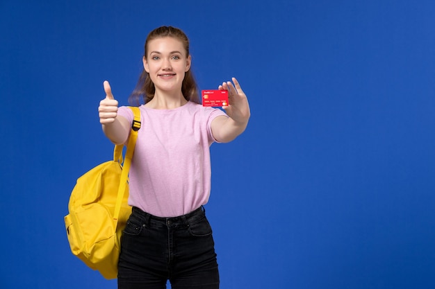 Front view of young female in pink t-shirt wearing yellow backpack holding plastic red card smiling on blue wall