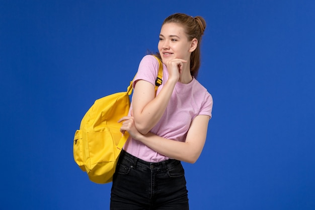 Front view of young female in pink t-shirt wearing yellow backpack posing on blue wall