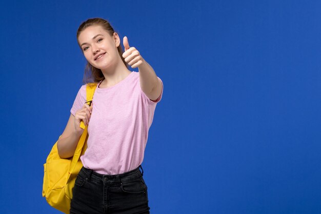 Front view of young female in pink t-shirt wearing yellow backpack posing smiling on the light-blue wall