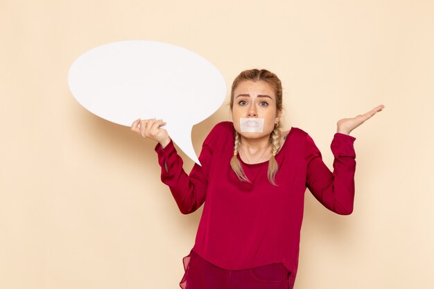 Front view young female in red shirt with tied mouth holding white huge sign on the cream space female cloth  photo violence domestic