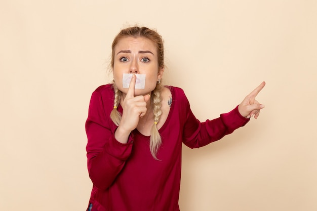 Front view young female in red shirt with tied mouth showing silence sign on the cream space female cloth 