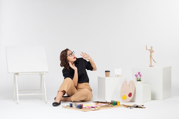 Front view young female sitting inside room with easel and paints on white background