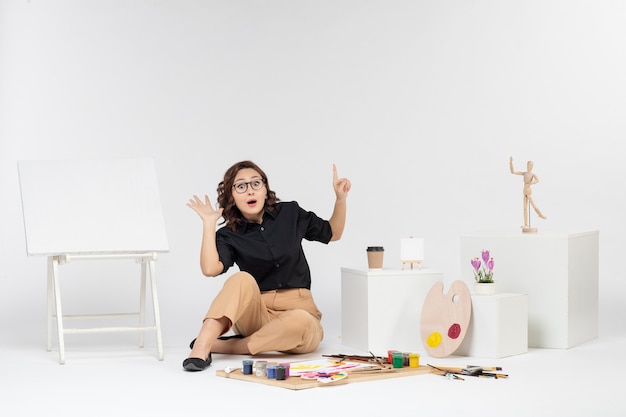 Front view young female sitting inside room with easel and paints on white desk color art draw picture painter artist drawing