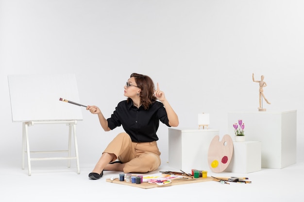 Front view young female sitting inside room with paints and easel on white background