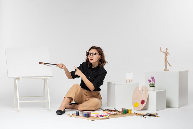 Front view young female sitting inside room with paints and easel on white background