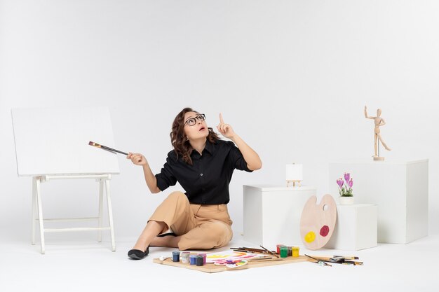 Front view young female sitting inside room with paints and easel on white background