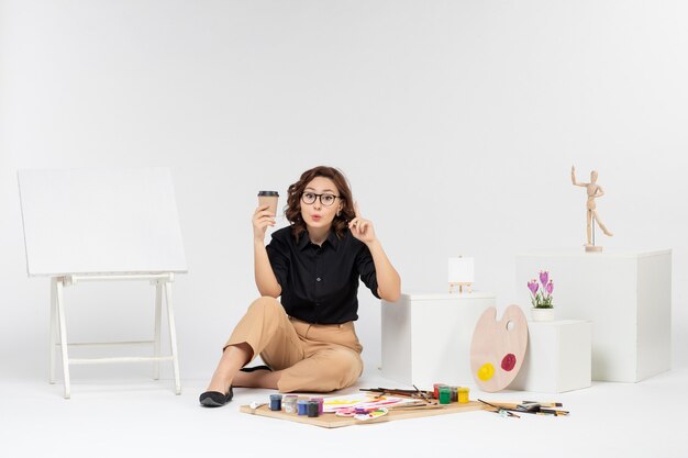 Front view young female sitting inside room with paints and easel on white background