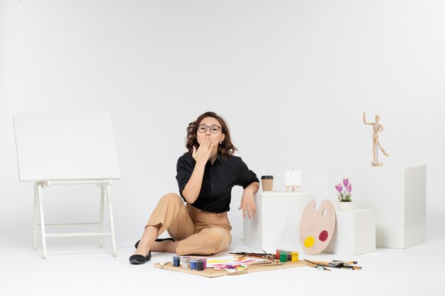 Front view young female sitting inside room with paints tassel and easel on white background